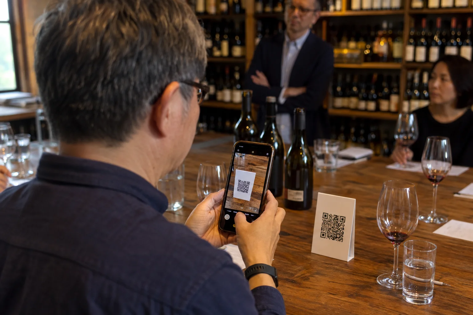 品酒會客人拿手機掃描木桌上的 QR Code 桌牌，桌邊備有品飲杯、飲水與筆記